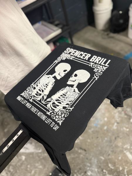 White screen-printed skeleton band-merch design on a black tee, on press at PrintForgeInk