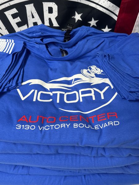 Stack of royal blue tees with a white and red Victory Auto Center back print and a flag print on the sleeve